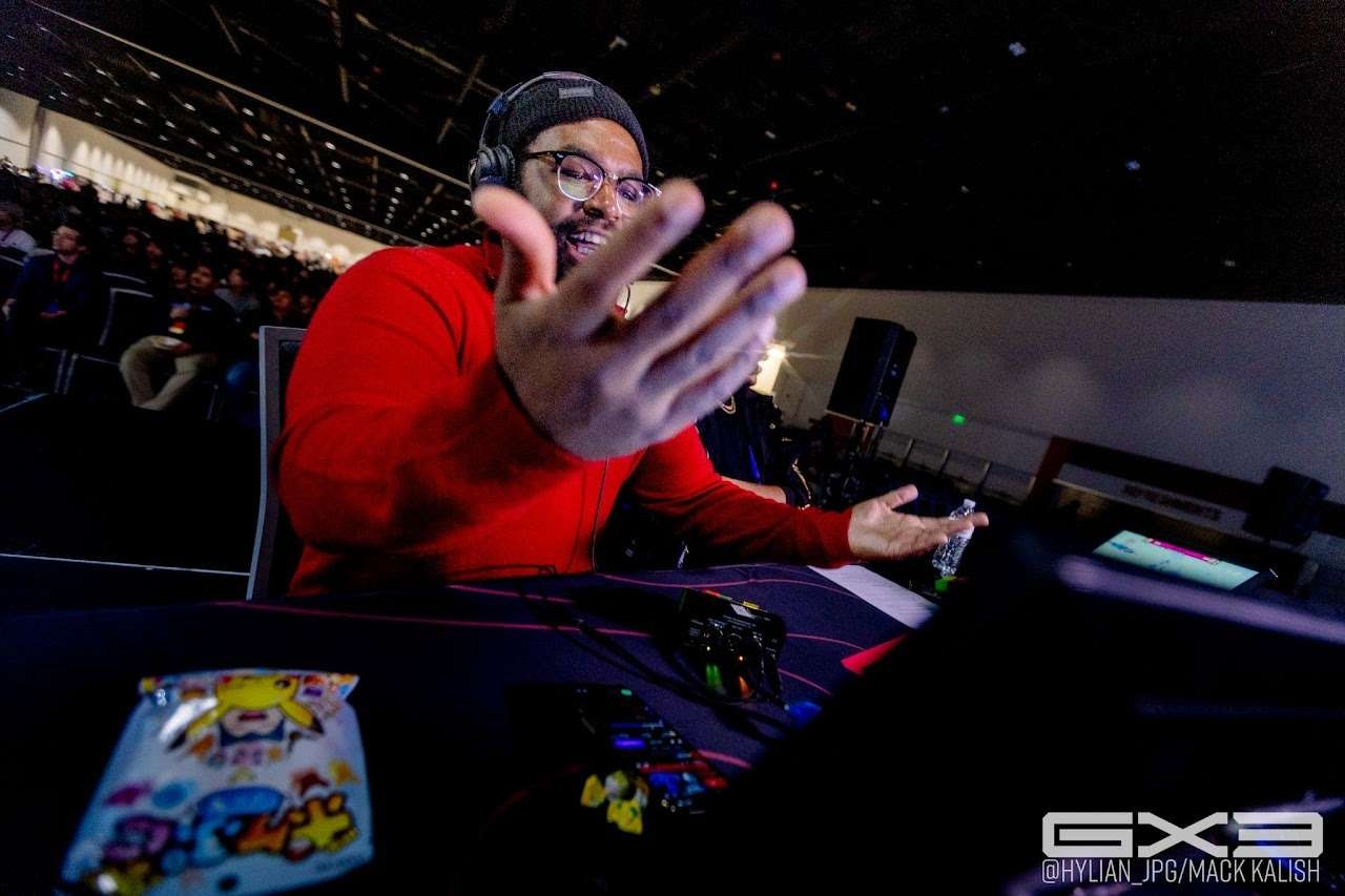 A live broadcast commentator reacts behind production equipment at GENESIS X3